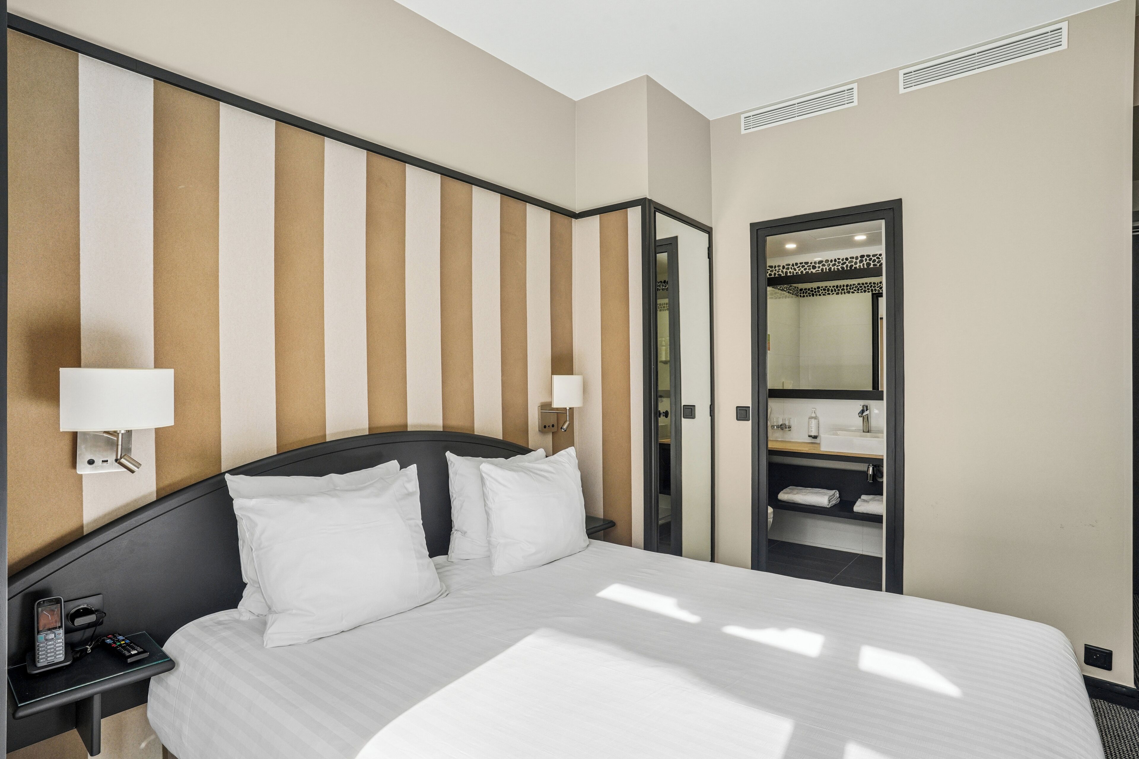 A hotel room with a bed, bedside tables, lamps, a mirror, and a bathroom visible through an open door.