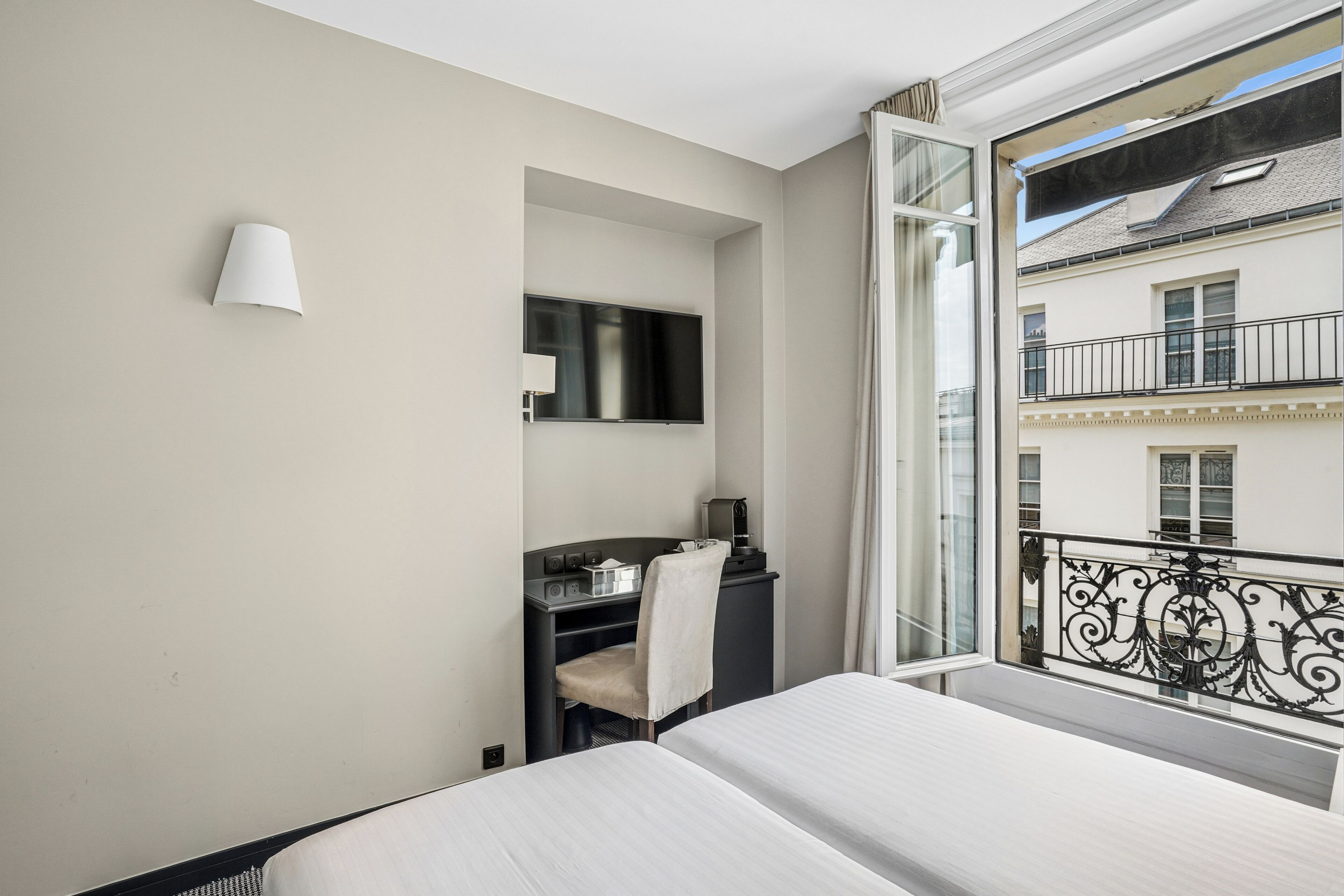 Habitación de hotel con cama, escritorio con silla, televisión y balcón con barandilla de hierro forjado.