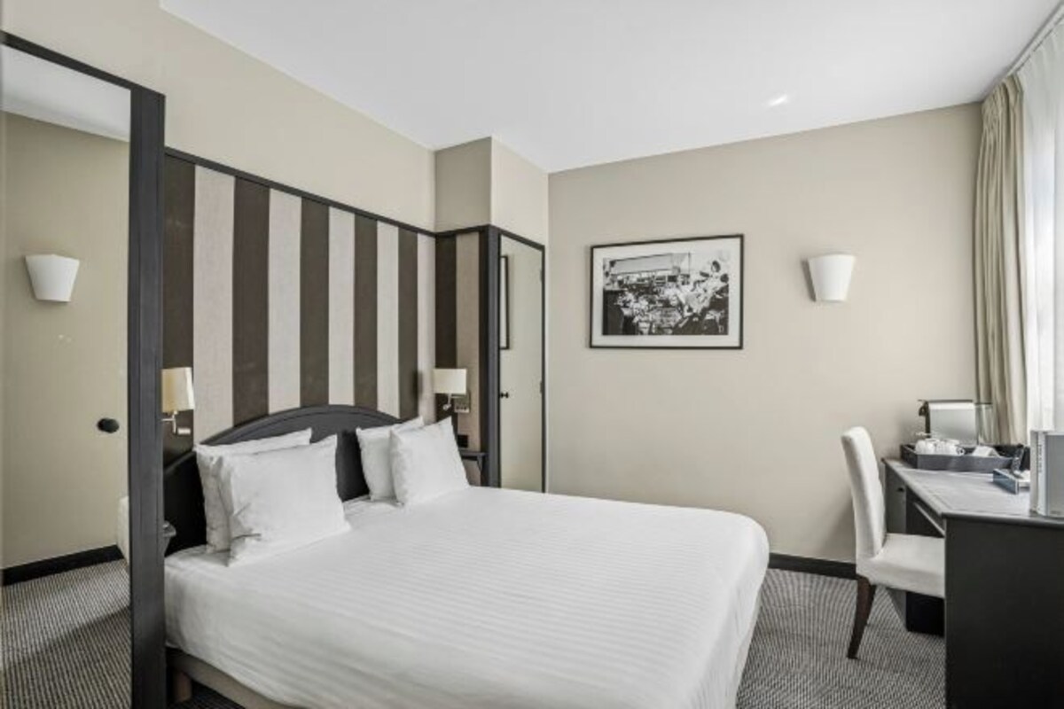A hotel room with a large bed, a desk, two chairs, a framed picture on the wall, and two wall-mounted lamps.