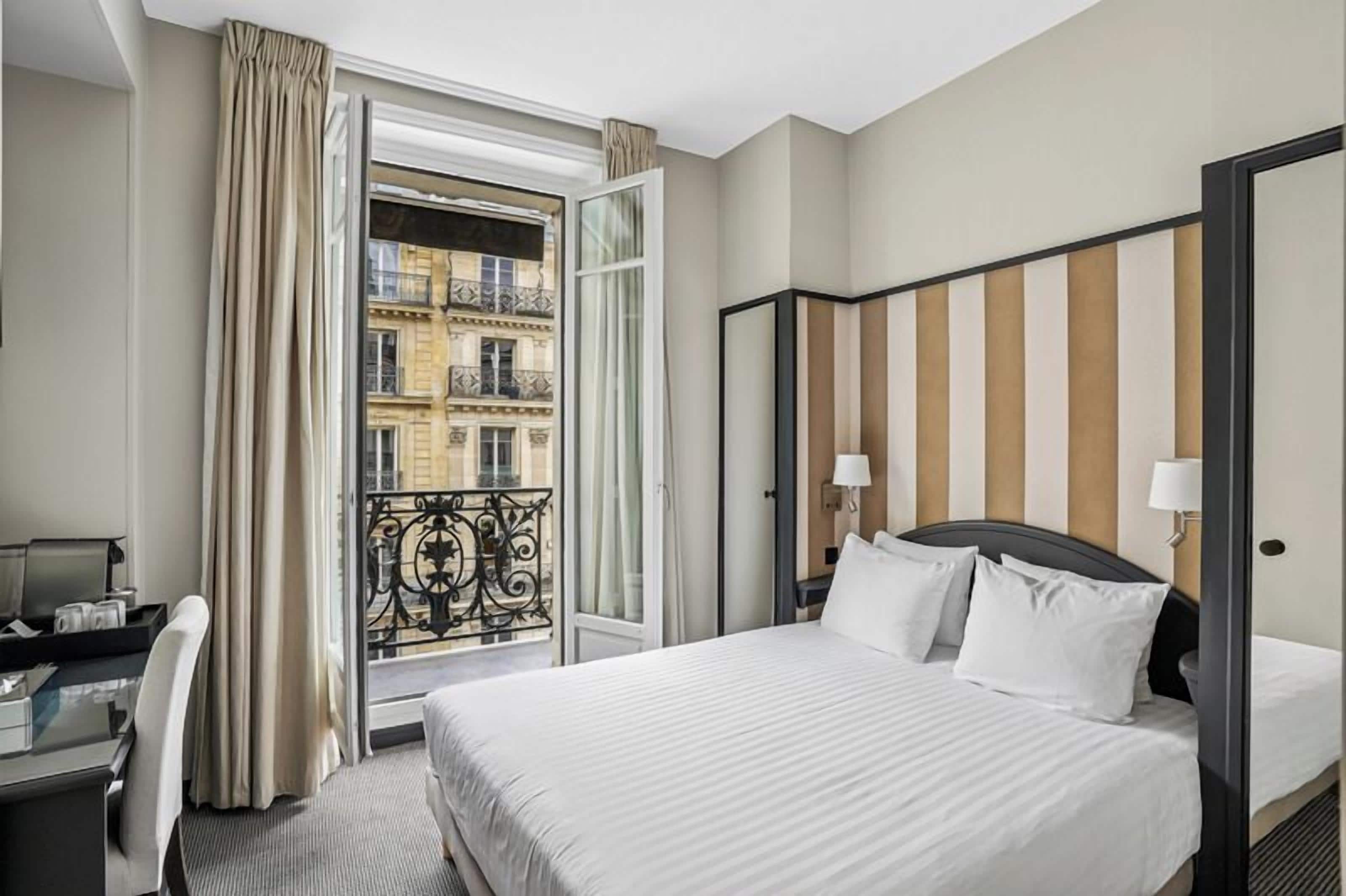 A hotel room with a bed, a desk, a chair, a window with curtains, and a view of buildings.