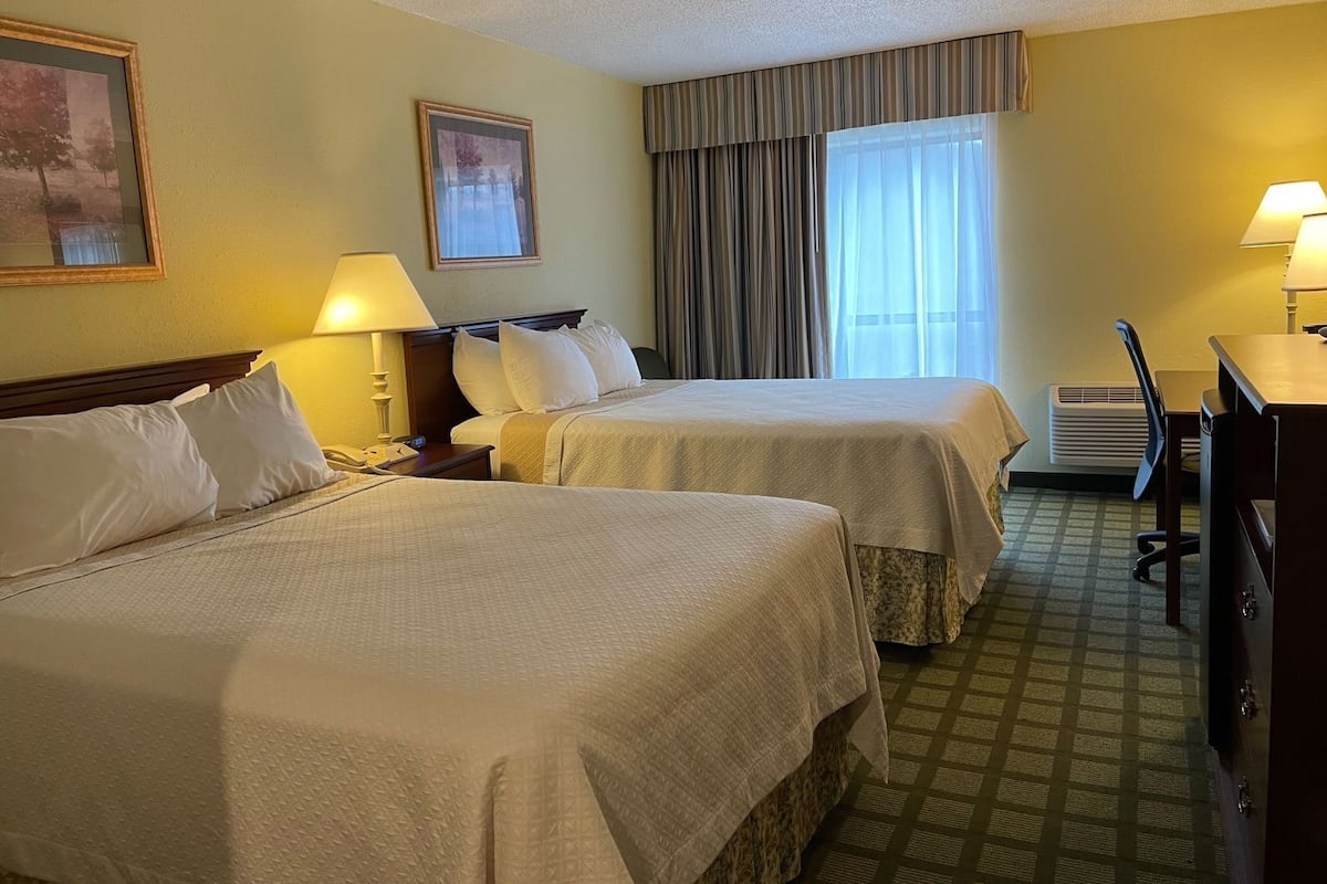 A hotel room with two beds, a desk, a chair, a lamp, a painting, and a window with curtains.