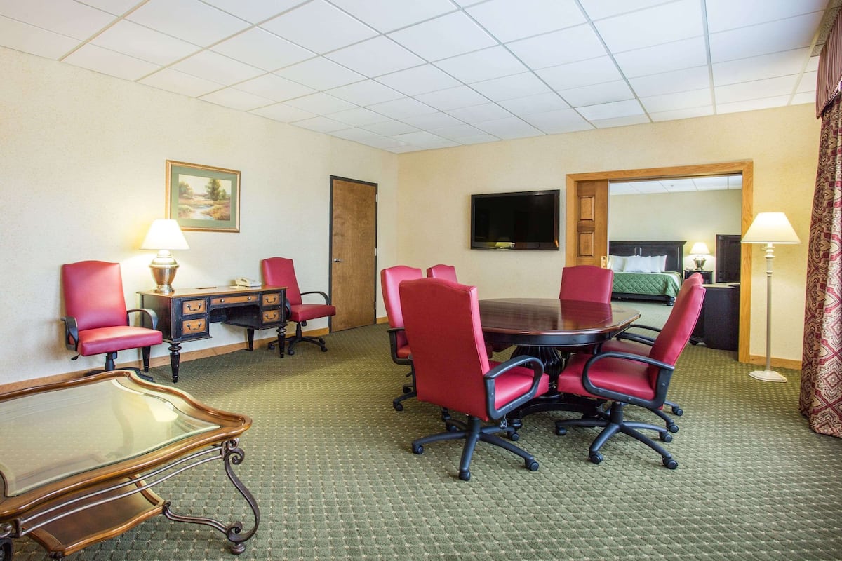 A hotel conference room with a large table, red chairs, a desk with a lamp, a TV on the wall, and a mirror.