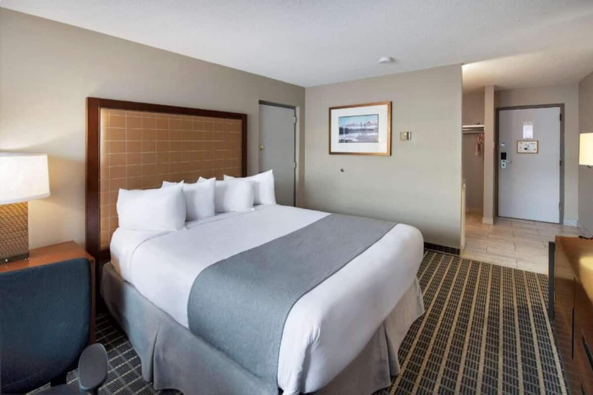 Habitación de hotel con una cama grande, un escritorio, una silla, un cuadro en la pared y una puerta que da a otra habitación.