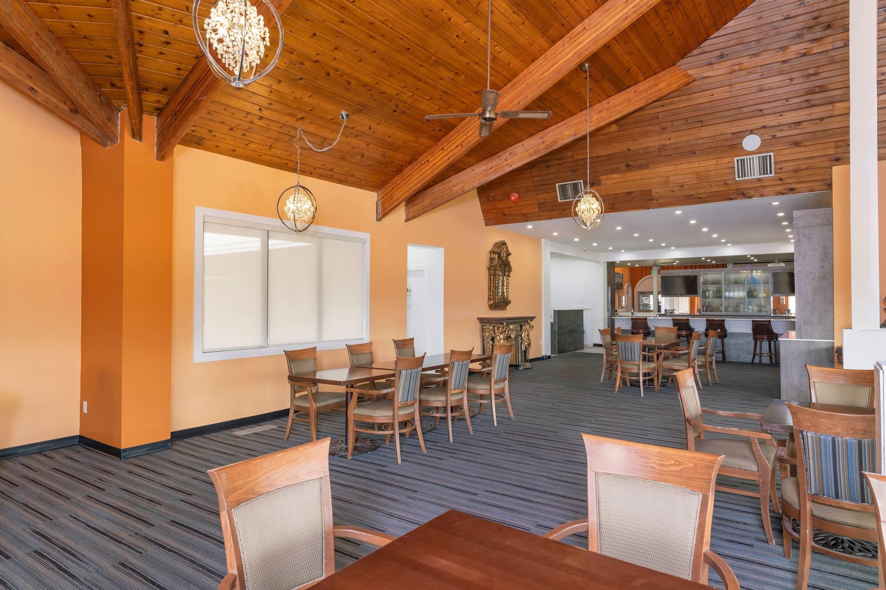 A spacious dining area with wooden tables and chairs, a high ceiling with exposed beams, and a kitchen in the background.