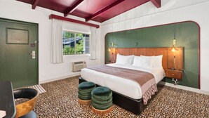 A bedroom with a bed, two ottomans, a nightstand, a lamp, a window with curtains, and a door.