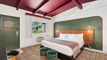 A bedroom with a bed, two ottomans, a nightstand, a lamp, a window with curtains, and a door.