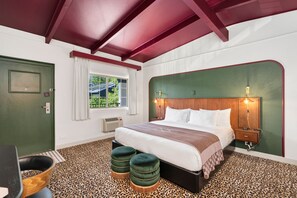 A bedroom with a bed, two ottomans, a nightstand, a lamp, a window with curtains, and a door.