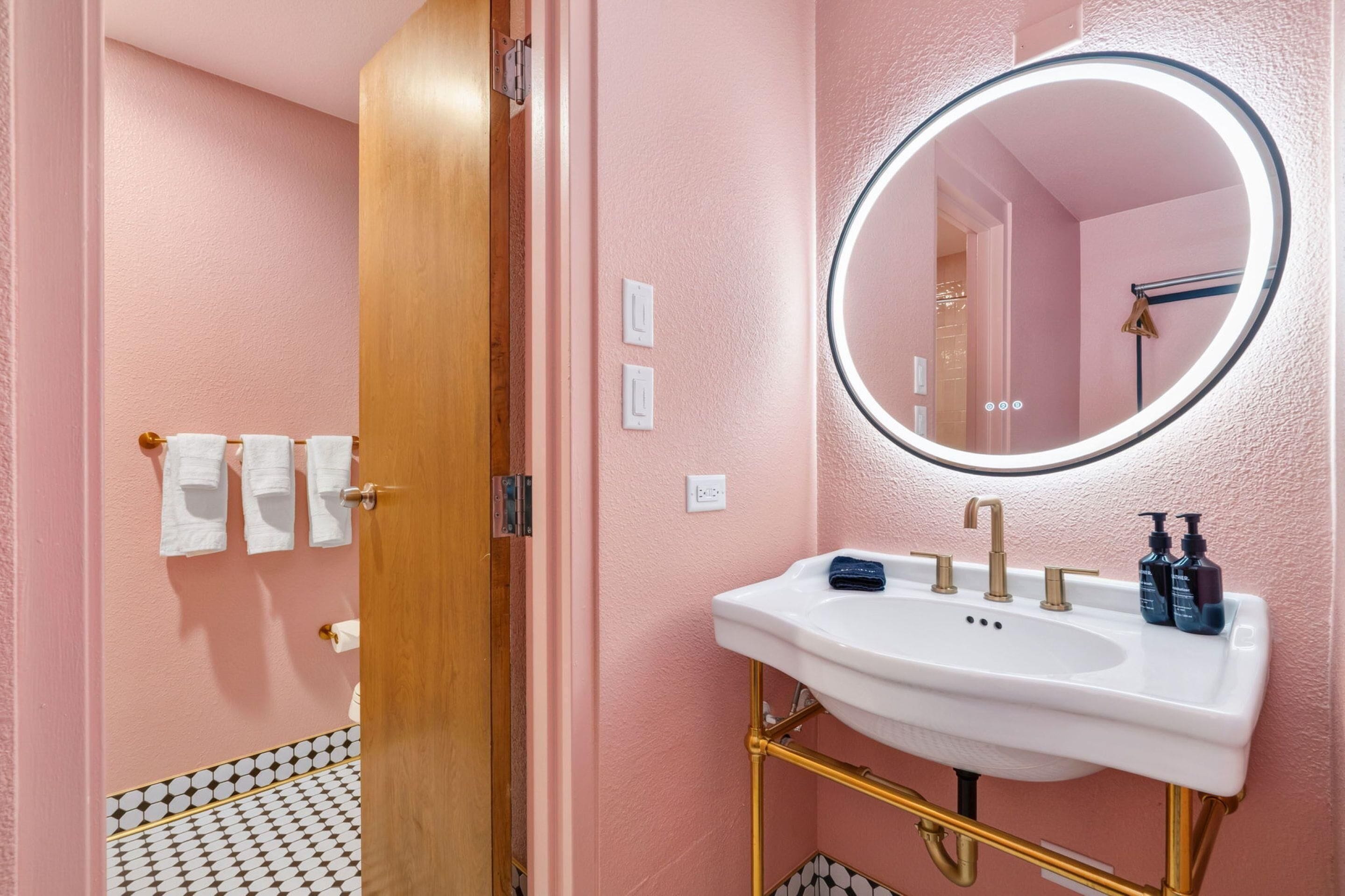A bathroom with a white sink, gold fixtures, a round mirror, and a pink wall.