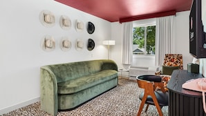 A room with a green sofa, a patterned carpet, a flat-screen TV, a desk with a telephone, and a window with curtains.