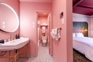 Ein Badezimmer mit Waschbecken, Spiegel und Pflegeprodukten. Im Hintergrund sind Toilette und Duschbereich zu sehen.
