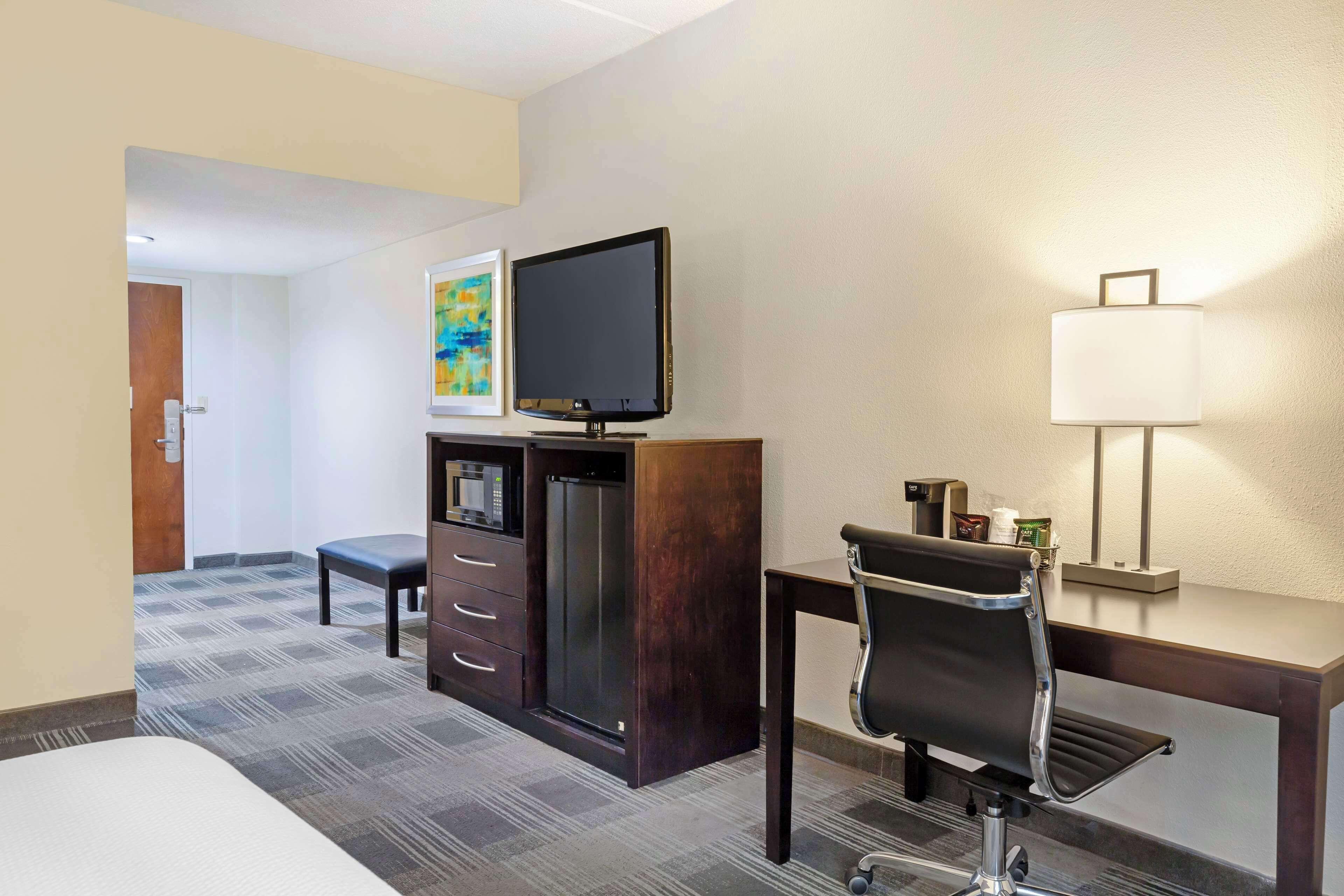 A hotel room with a bed, a desk with a chair, a television, a refrigerator, a microwave, a coffee maker, and a lamp.