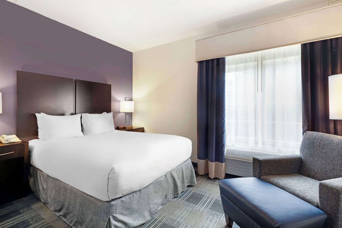 Ein Hotelzimmer mit einem großen Bett, einem Nachttisch mit Telefon, einem Sessel und einem Fenster mit Vorhängen.