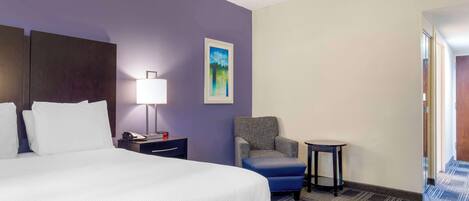 Habitación de hotel con una cama grande, una mesita de noche con lámpara, una silla y una pequeña mesa.