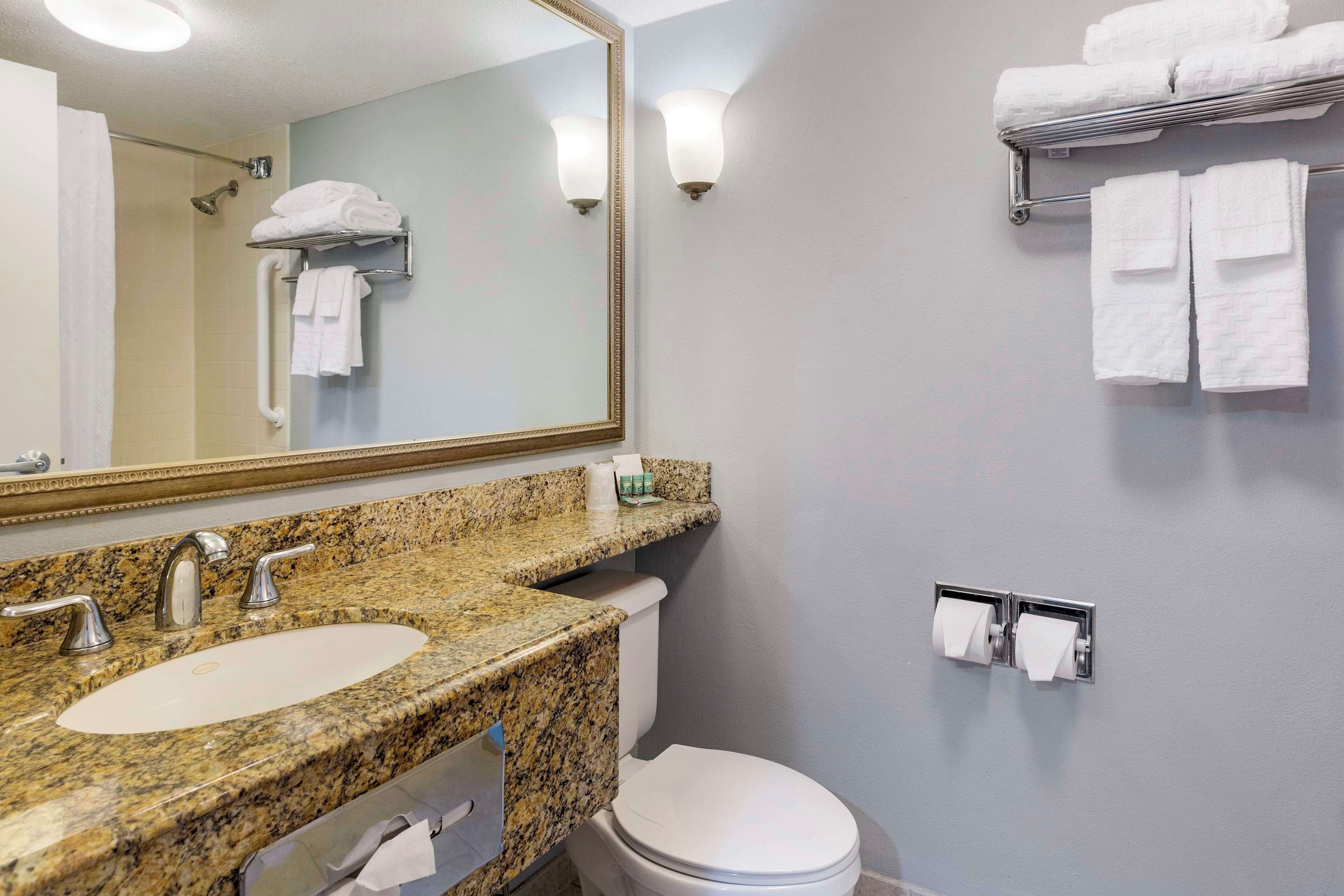 A bathroom with a granite countertop sink, a white toilet, a large mirror, and a towel rack with folded towels.