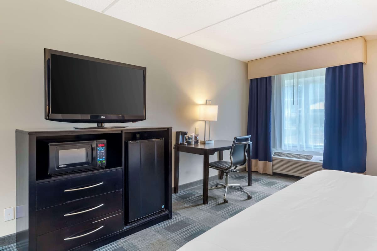Ein Hotelzimmer mit einem Bett, einem Schreibtisch mit Stuhl, einem Fernseher, einer Mikrowelle, einem Kühlschrank und einem Fenster mit Vorhängen.