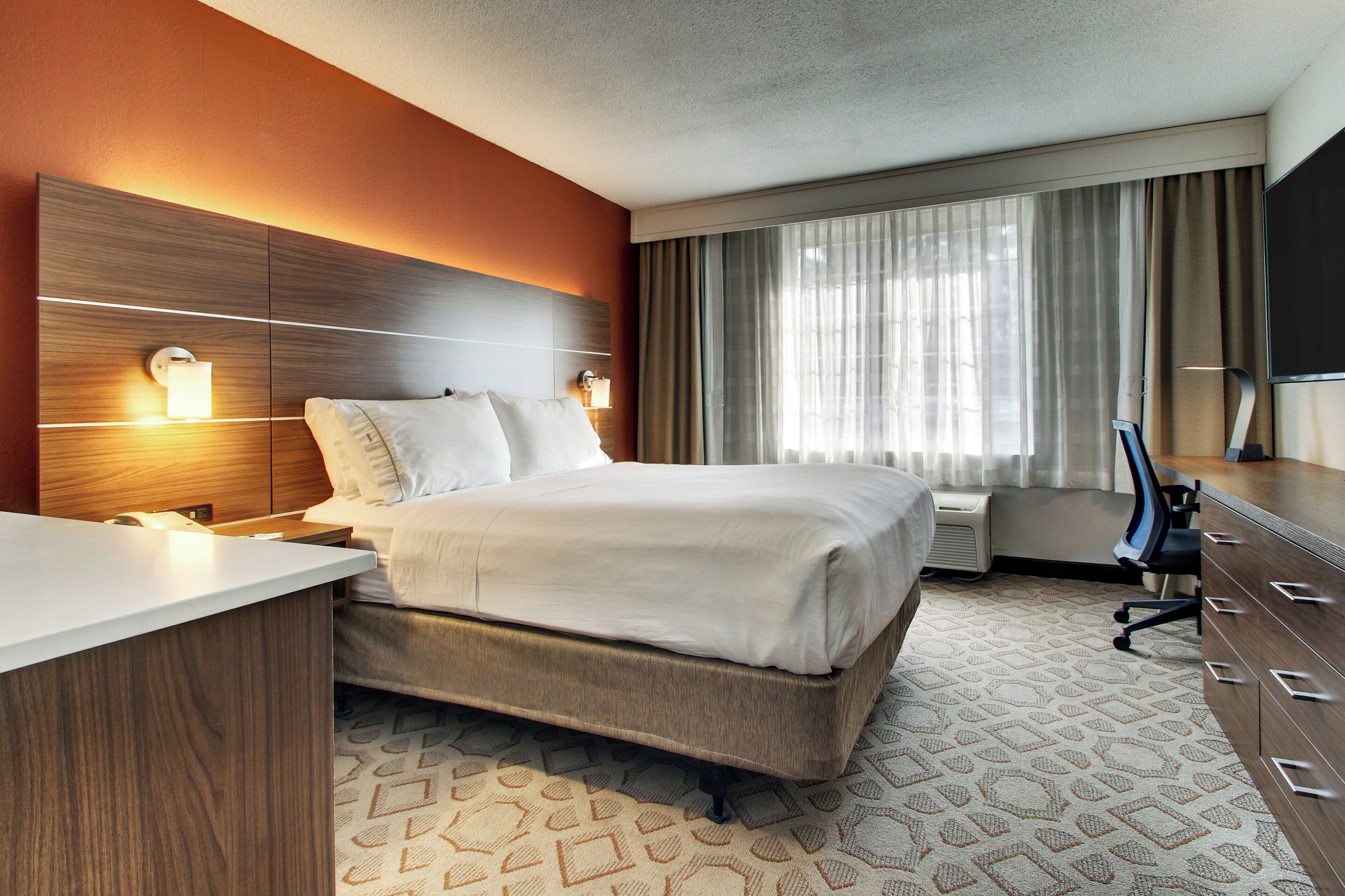 A hotel room with a bed, bedside tables, a desk, a chair, a TV, and a window with curtains.