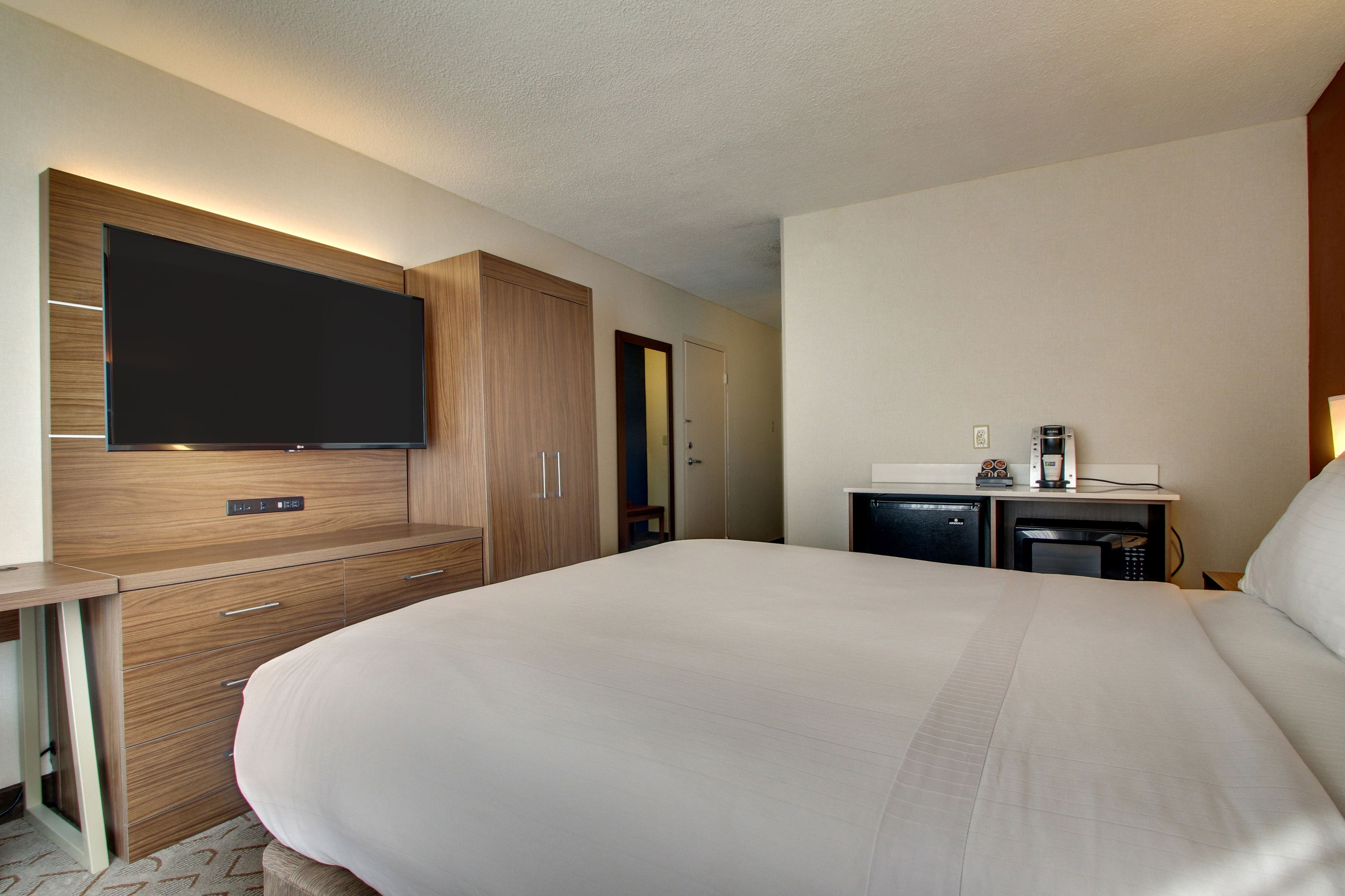 Una habitación de hotel con una cama grande, un televisor de pantalla plana montado en un mueble de madera, una pequeña kitchenette con una cafetera, y una puerta que da a otra habitación.