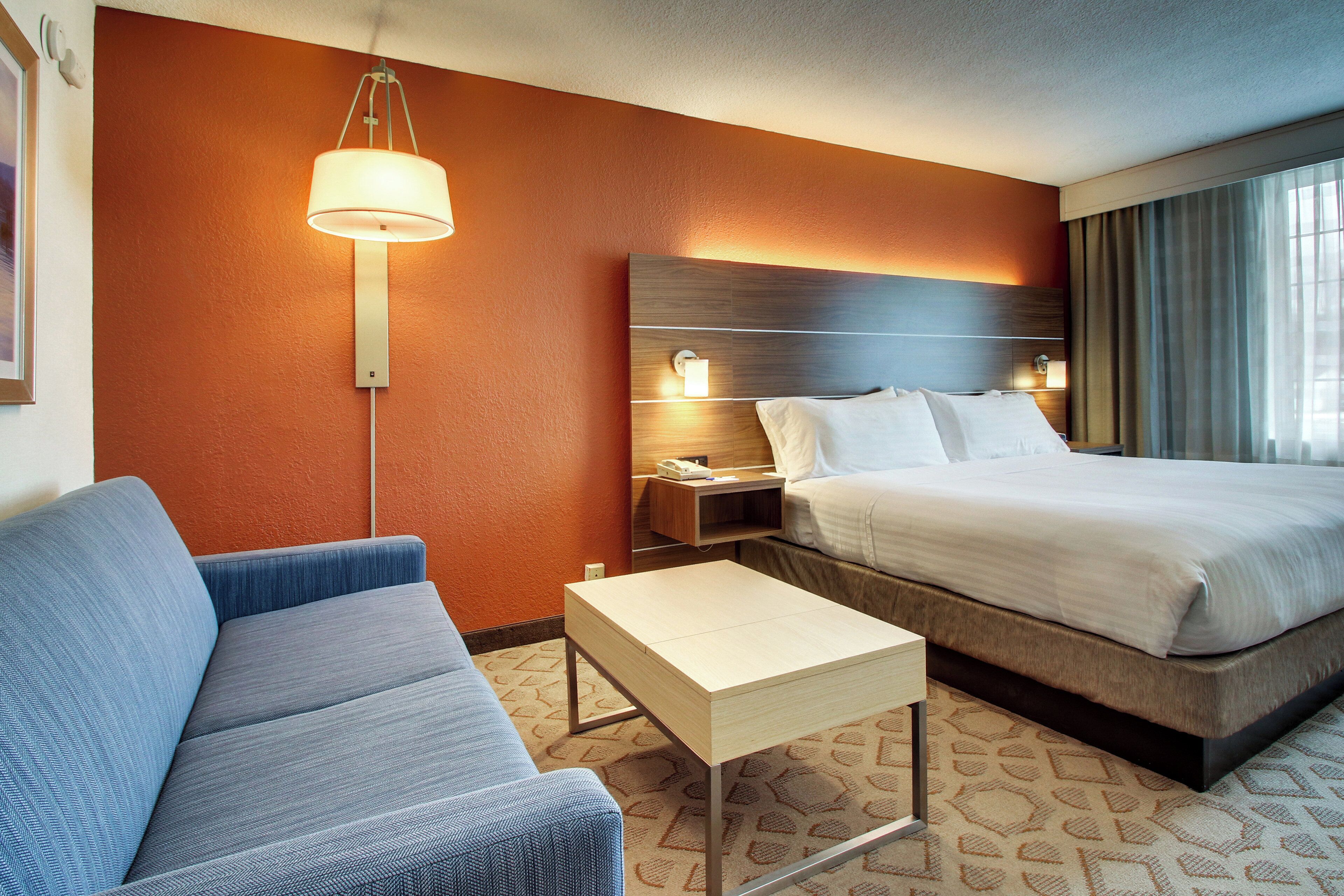 A hotel room with a bed, a sofa, a nightstand, a lamp, and a carpeted floor.
