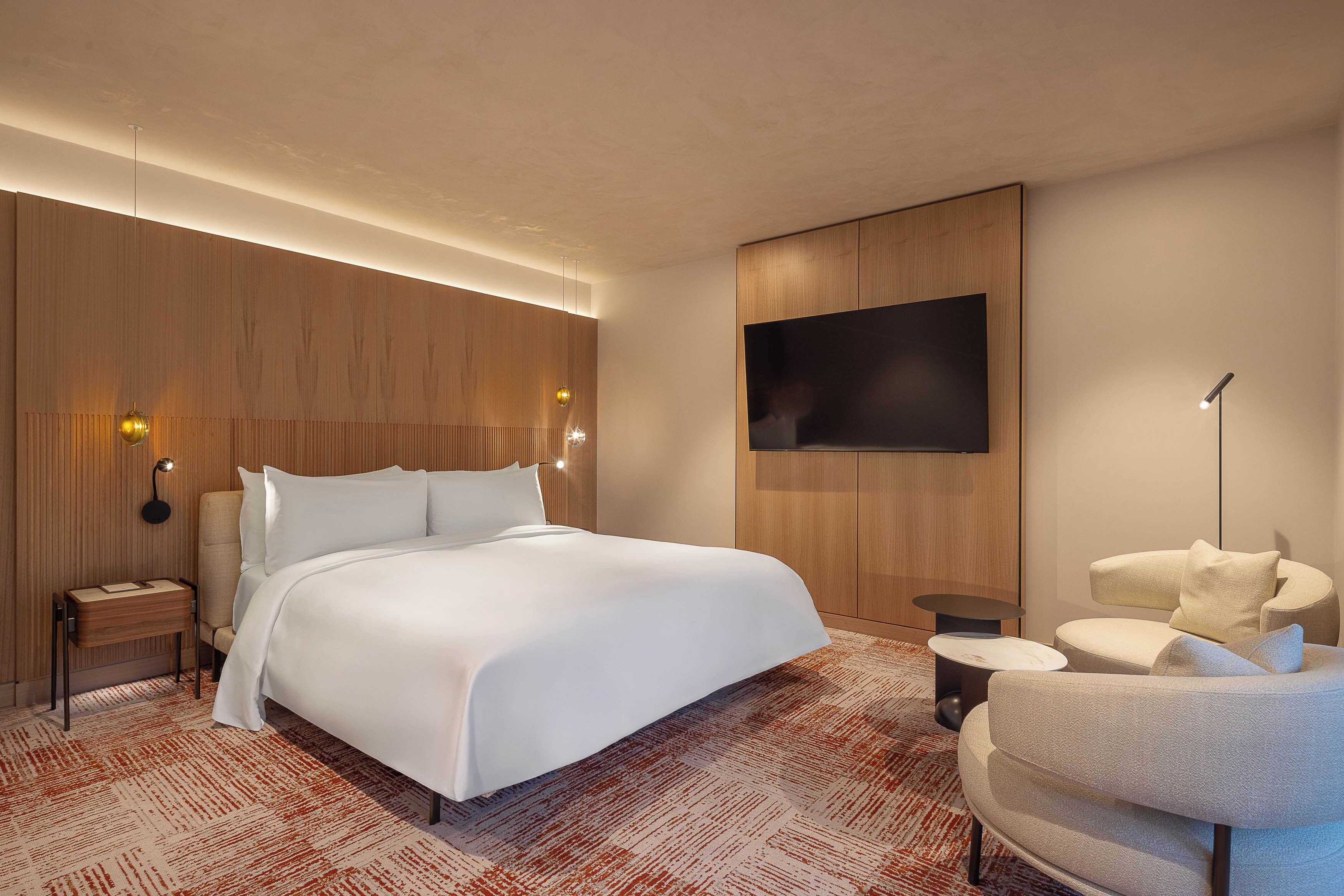 Quarto de hotel moderno com uma cama grande, televisão de tela plana na parede, mesa redonda pequena, sofá e mesa de cabeceira com luminária.