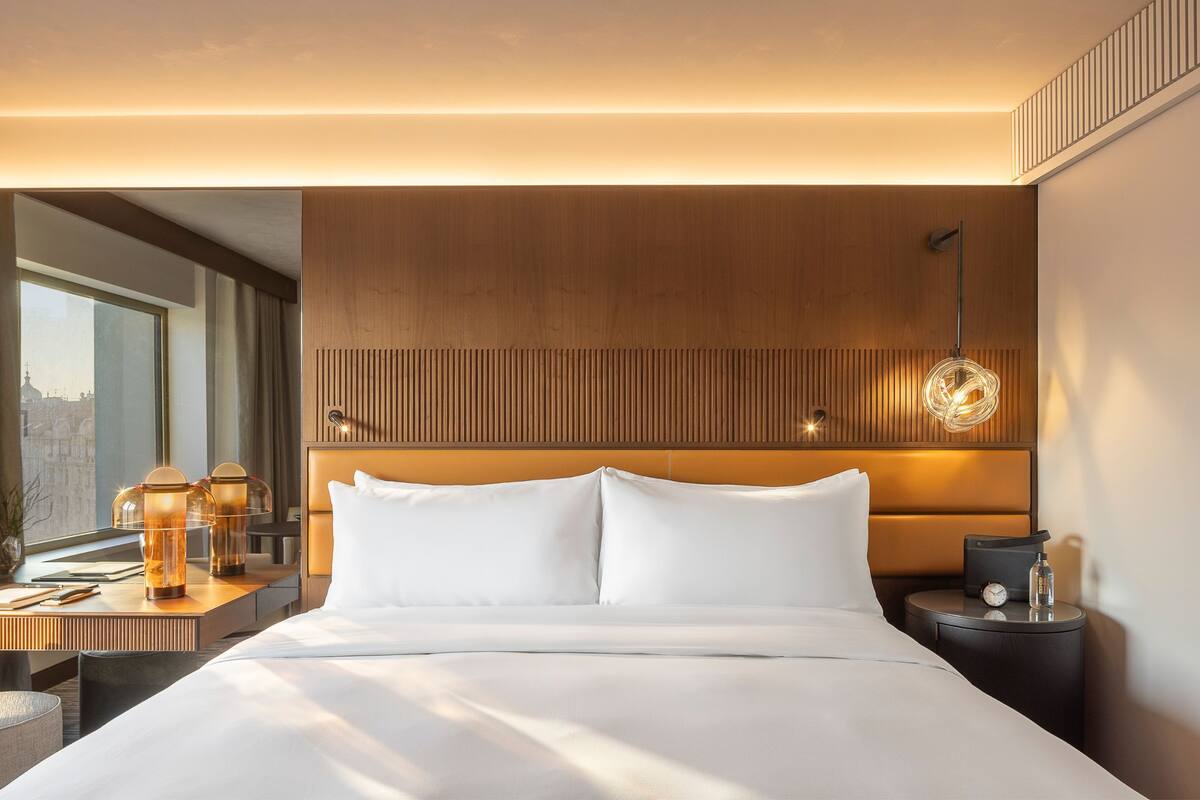 Habitación de hotel moderna con una cama grande, cabecera de madera, mesita de noche con lámpara y vistas a la ciudad.