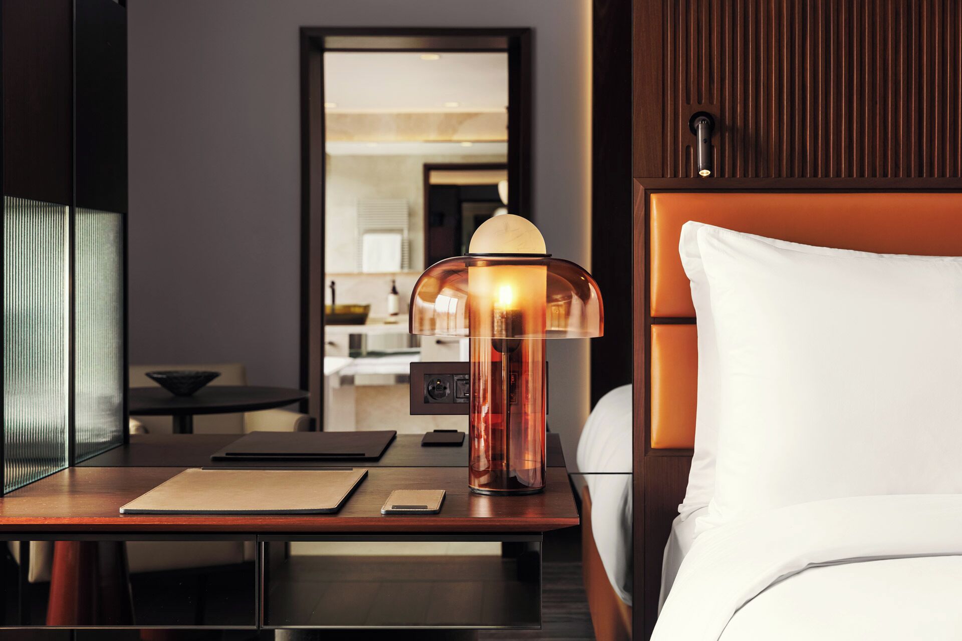 A hotel room with a bedside table, a lamp, a bed with white bedding, and a view of the bathroom and kitchen through an open door.
