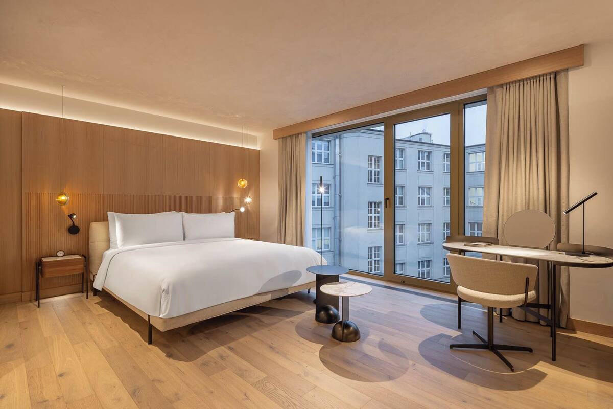 Habitación de hotel con una cama grande, un escritorio, una silla y vistas a edificios a través de amplios ventanales.