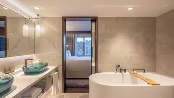 A modern bathroom with a glass sink, a large bathtub, and a view of a bedroom through a doorway.