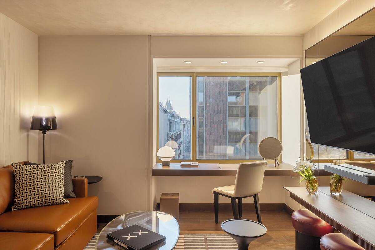 Una habitación de hotel moderna con sofá, una mesa de centro, escritorio con silla, televisor y ventana grande con vistas a la ciudad.