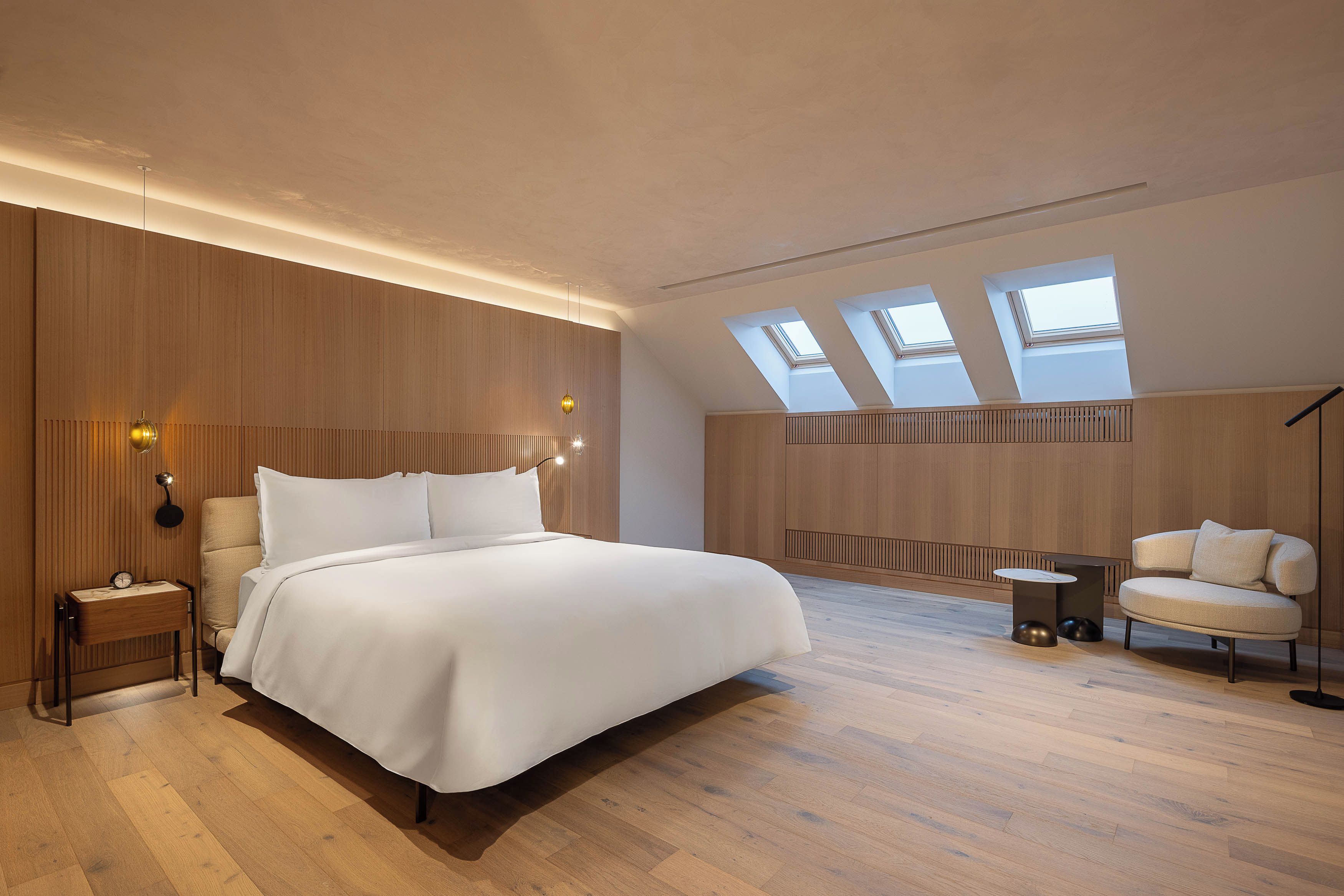Un dormitorio moderno con una cama grande, una mesita de noche, una lámpara, una silla y una pequeña mesa. Hay tragaluces en el techo.