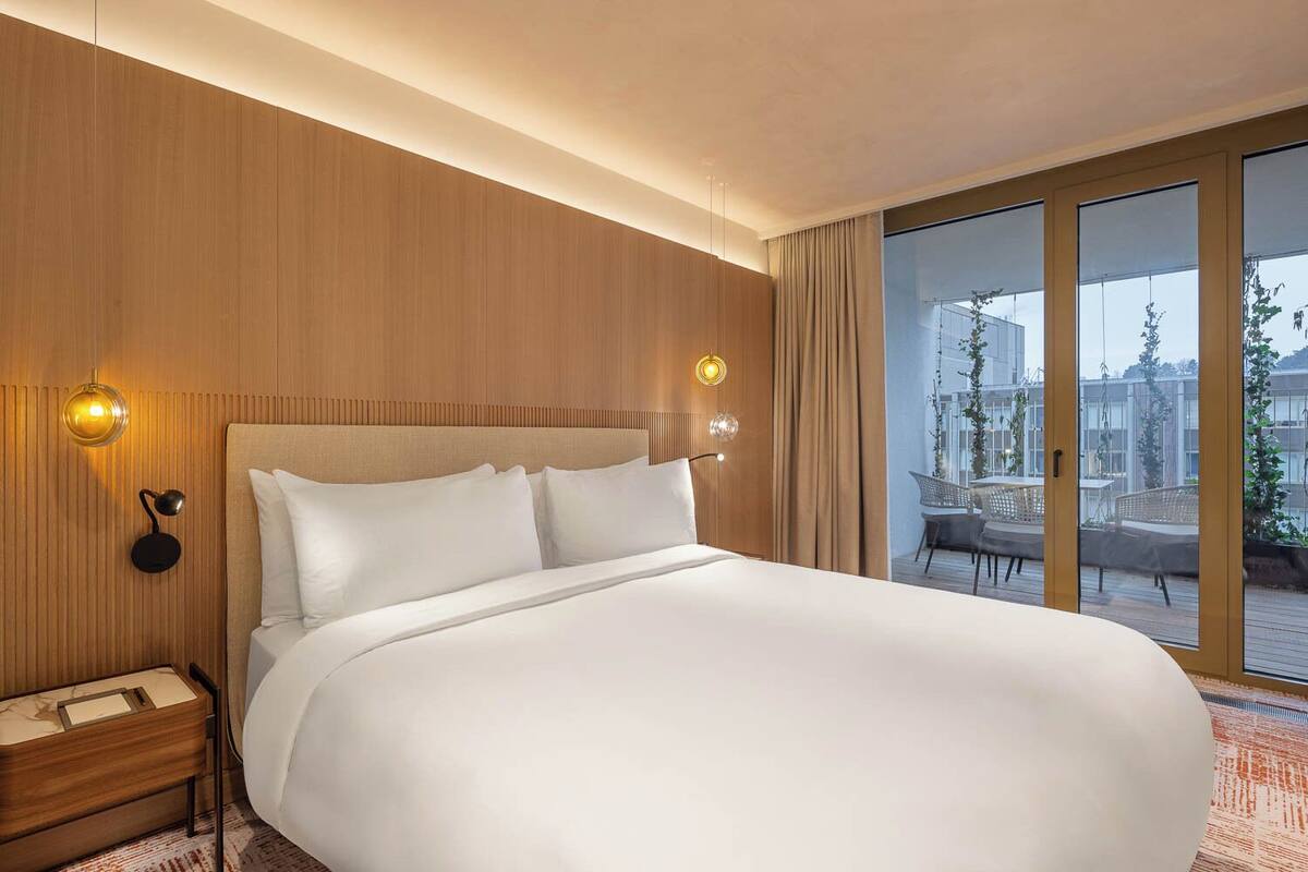 Una cama grande con sábanas blancas en una habitación con paneles de madera y vistas a un balcón con mobiliario exterior.