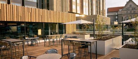 An outdoor seating area with tables and chairs, a wooden deck, and a modern building with large windows in the background.