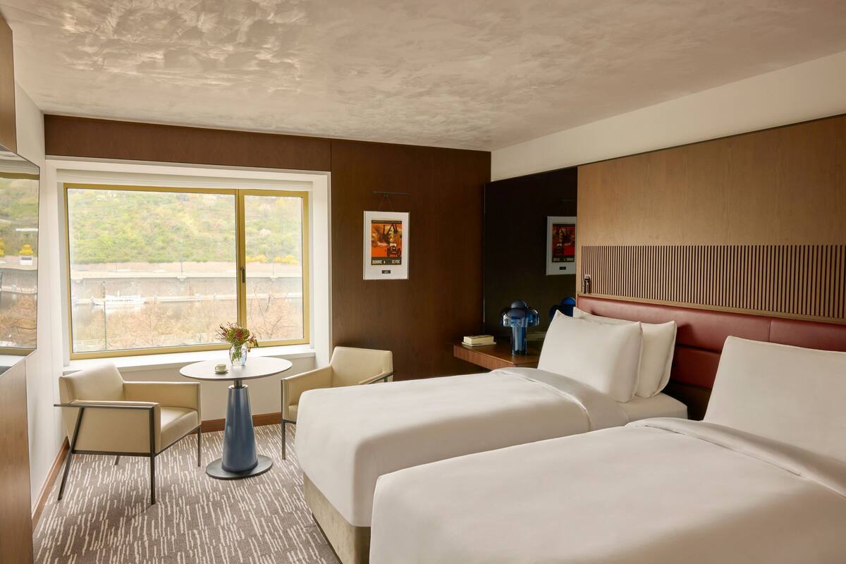 Habitación de hotel con dos camas, una mesita pequeña, una silla y un ventanal con vistas al exterior.