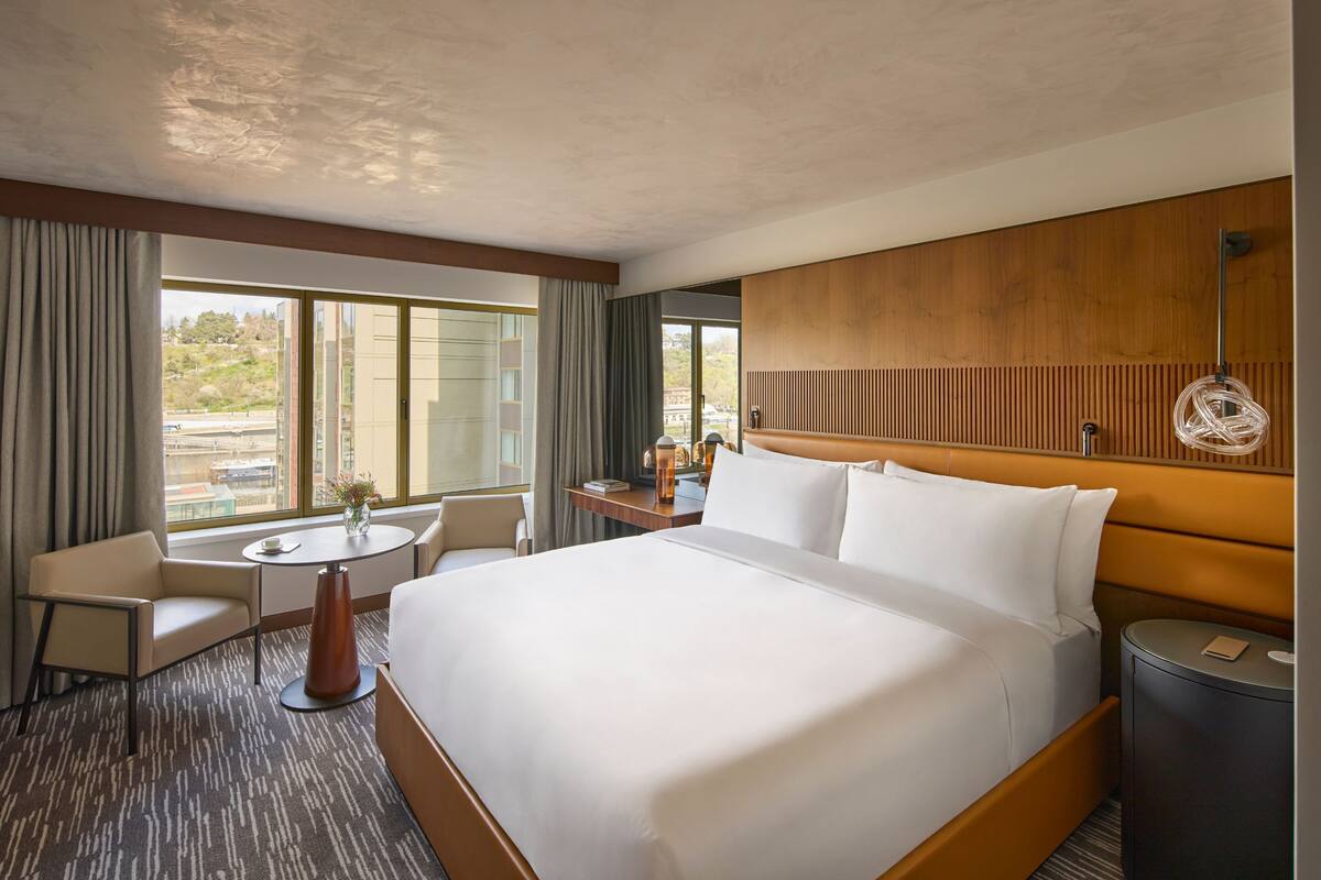 Habitación de hotel con una cama grande, un escritorio, una silla y vistas de la ciudad a través de amplios ventanales.