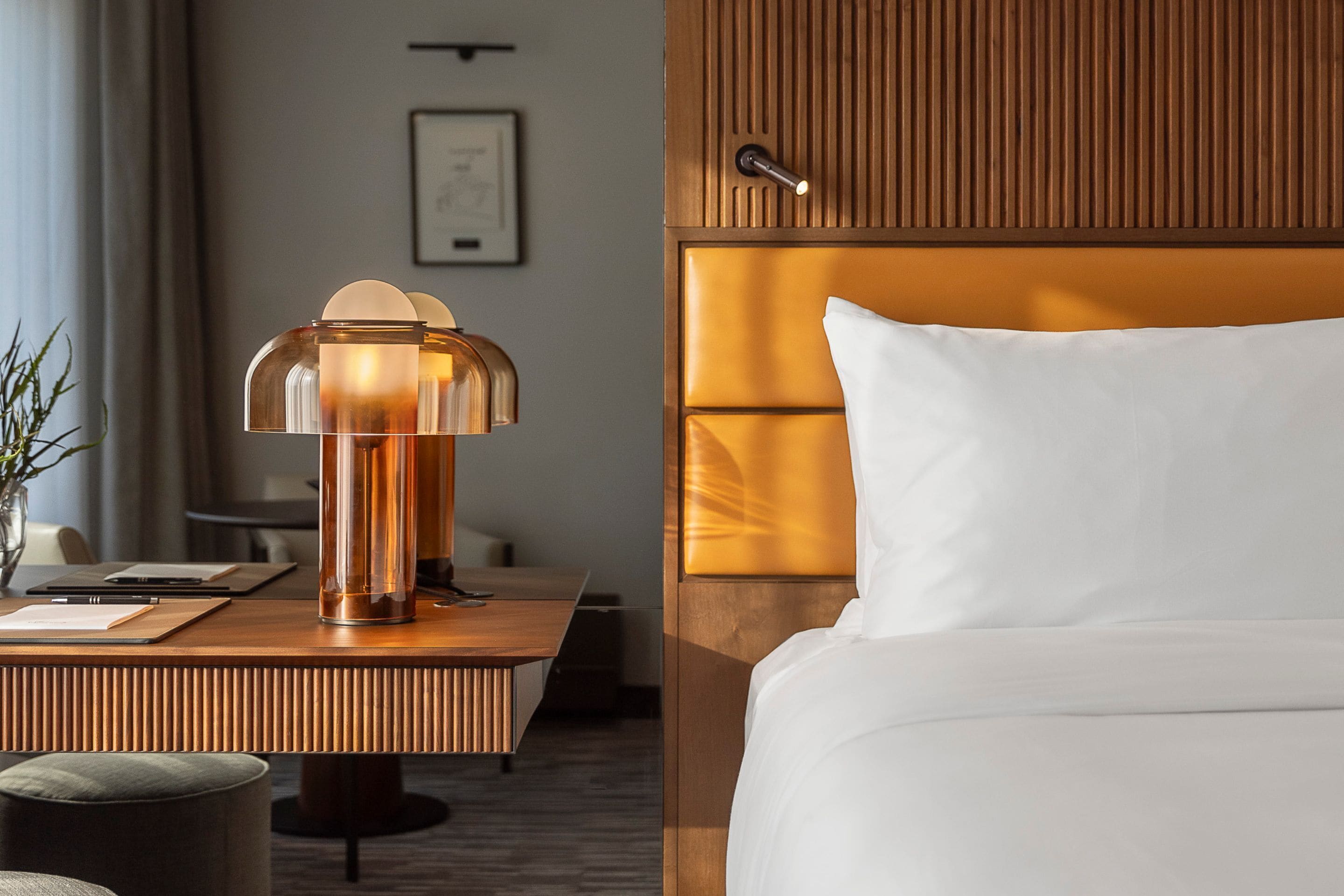A hotel room with a wooden headboard, a bedside table with a lamp, a bed with white linens, and a framed picture on the wall.