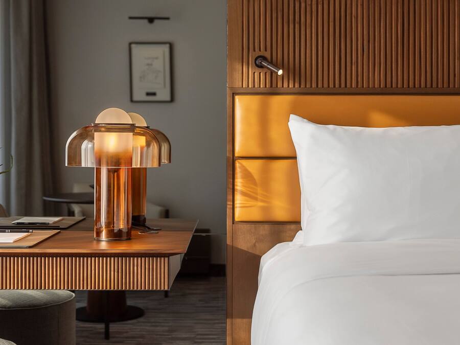 A hotel room with a wooden headboard, a bedside table with a lamp, a bed with white linens, and a framed picture on the wall.