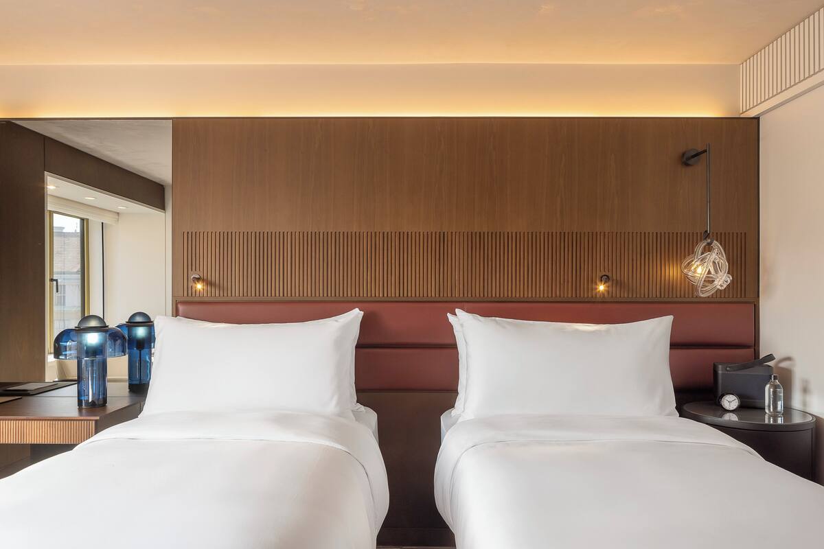 Habitación de hotel con dos camas, cabecera de madera, mesita de noche con lámpara y ventana con persianas.