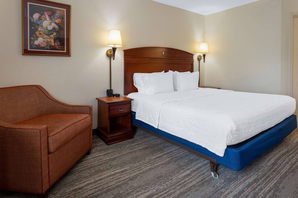 A hotel room with a bed, a nightstand, a lamp, a framed picture on the wall, and a brown armchair.