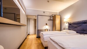 Une chambre d’hôtel avec deux lits, un plancher en bois, un miroir, une lampe fixée au mur et une porte donnant sur une autre pièce.