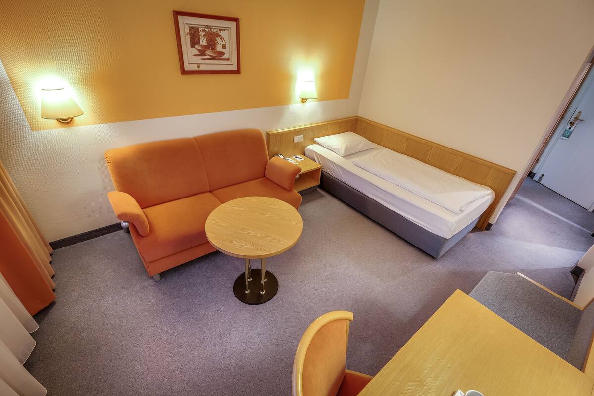 Une chambre d’hôtel compacte comprenant un canapé, une petite table ronde, un lit simple et deux lampes fixées au mur.