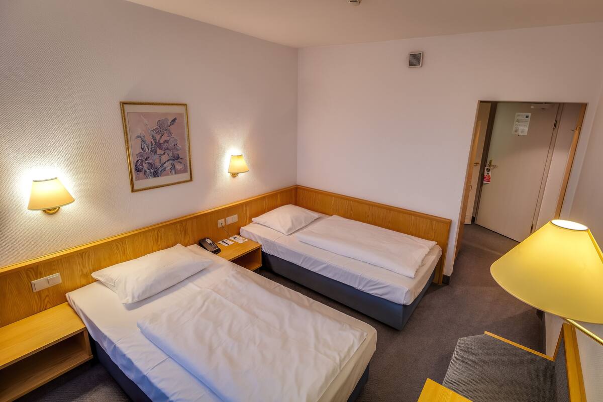 Une chambre d’hôtel avec deux lits simples, une tête de lit en bois, une table de chevet avec une lampe, une œuvre d’art florale encadrée au mur et une porte avec une serrure.