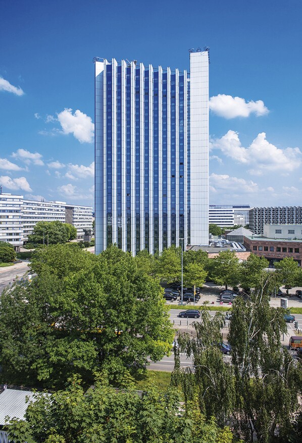 Een modern hoog gebouw met een glazen gevel, omgeven door bomen en een parkeerterrein.