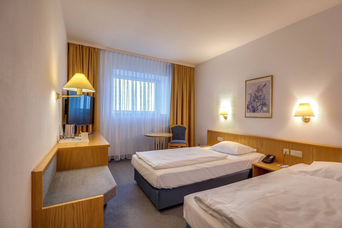 Une chambre d’hôtel avec deux lits, un bureau, une chaise, une petite table et une fenêtre avec des rideaux.