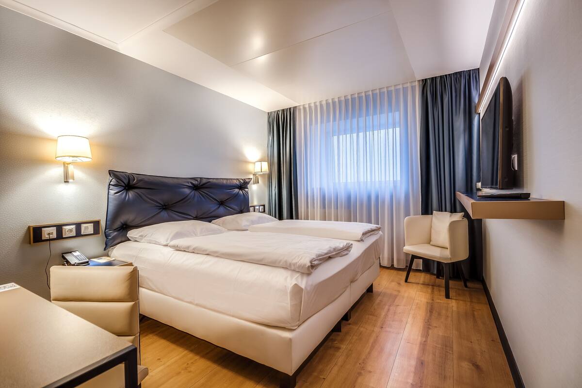 Et hotelværelse med en stor seng, sengeborde med lamper, et skrivebord, en stol, et fjernsyn og et vindue med gardiner.
