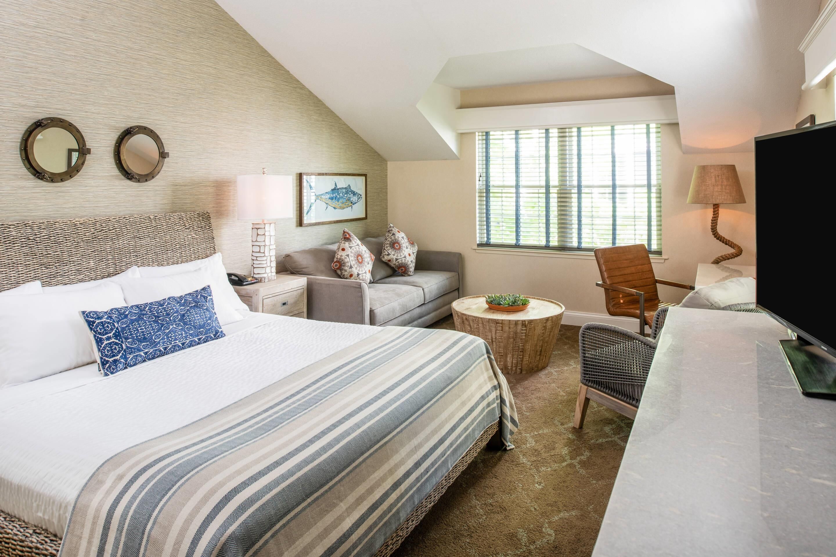 A hotel room with a large bed, a sofa, a small round table, a chair, a TV, and a window with blinds.