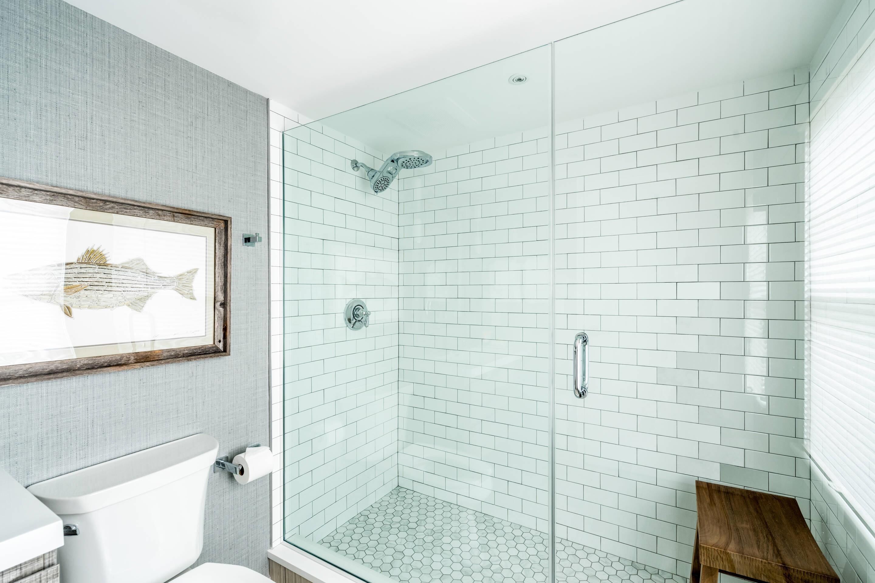 A modern bathroom with a glass shower enclosure, white hexagonal tiles, a wooden stool, and a framed fish artwork on the wall.