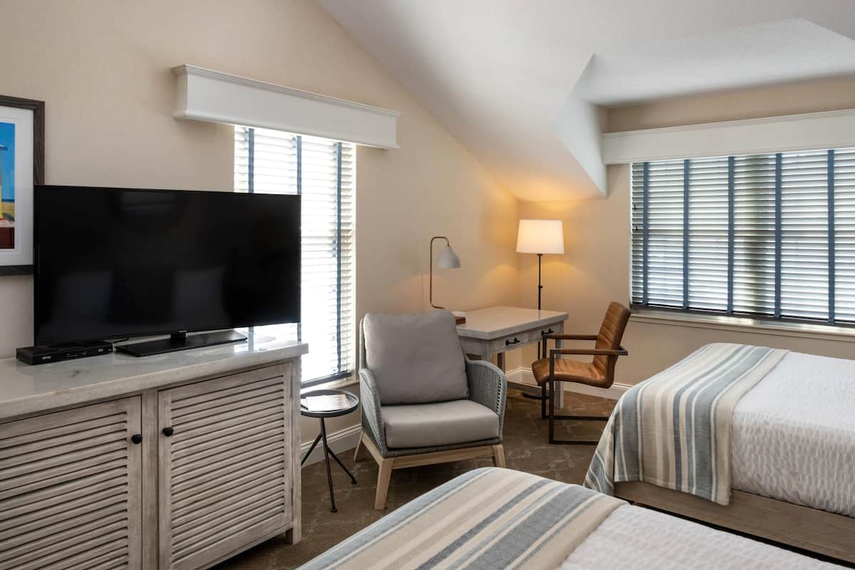 A hotel room with a flat-screen TV, a desk, two chairs, a lamp, and two beds.