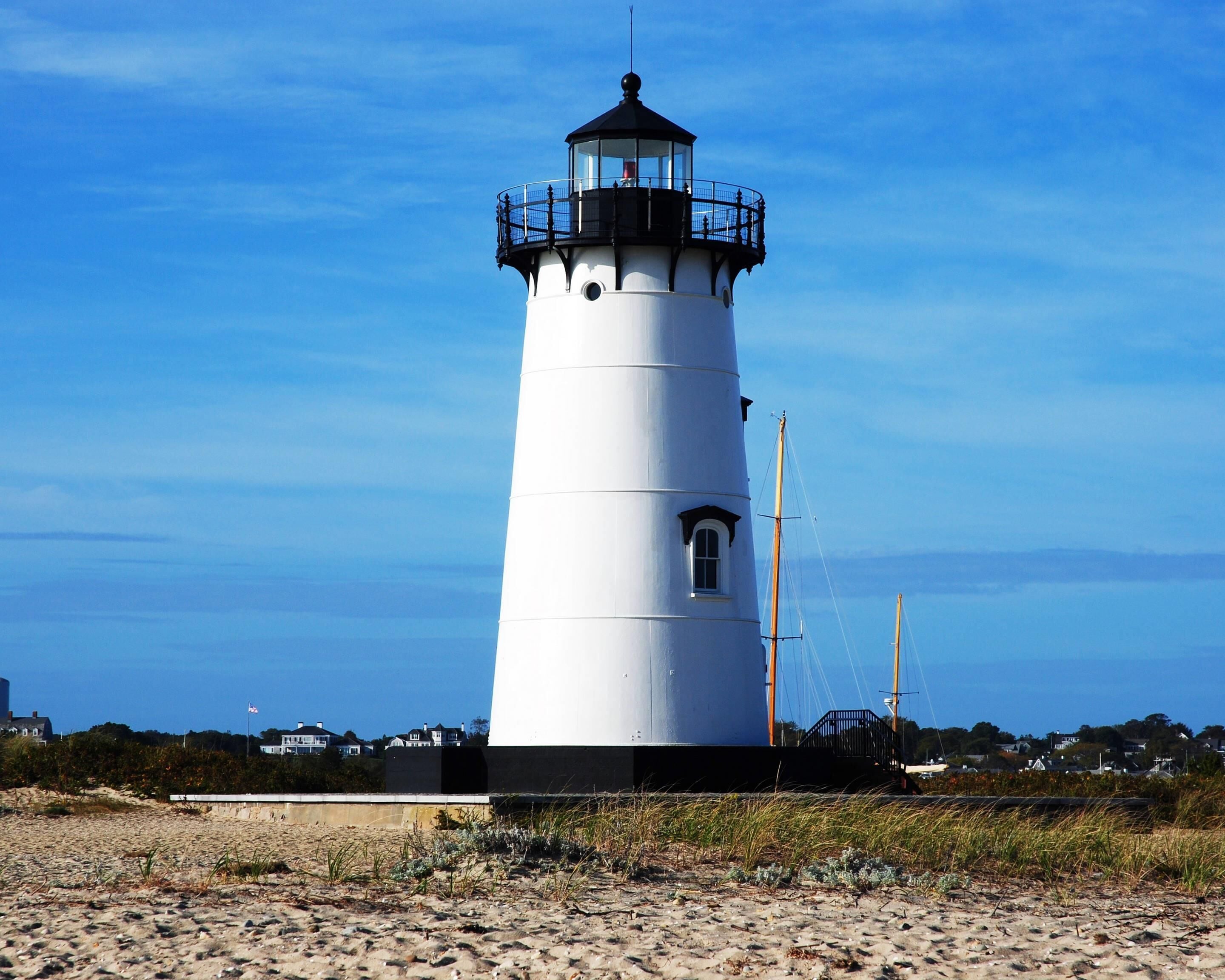 Un phare avec une partie supérieure noire et un corps blanc, situé sur une plage de sable, sous un ciel bleu dégagé.