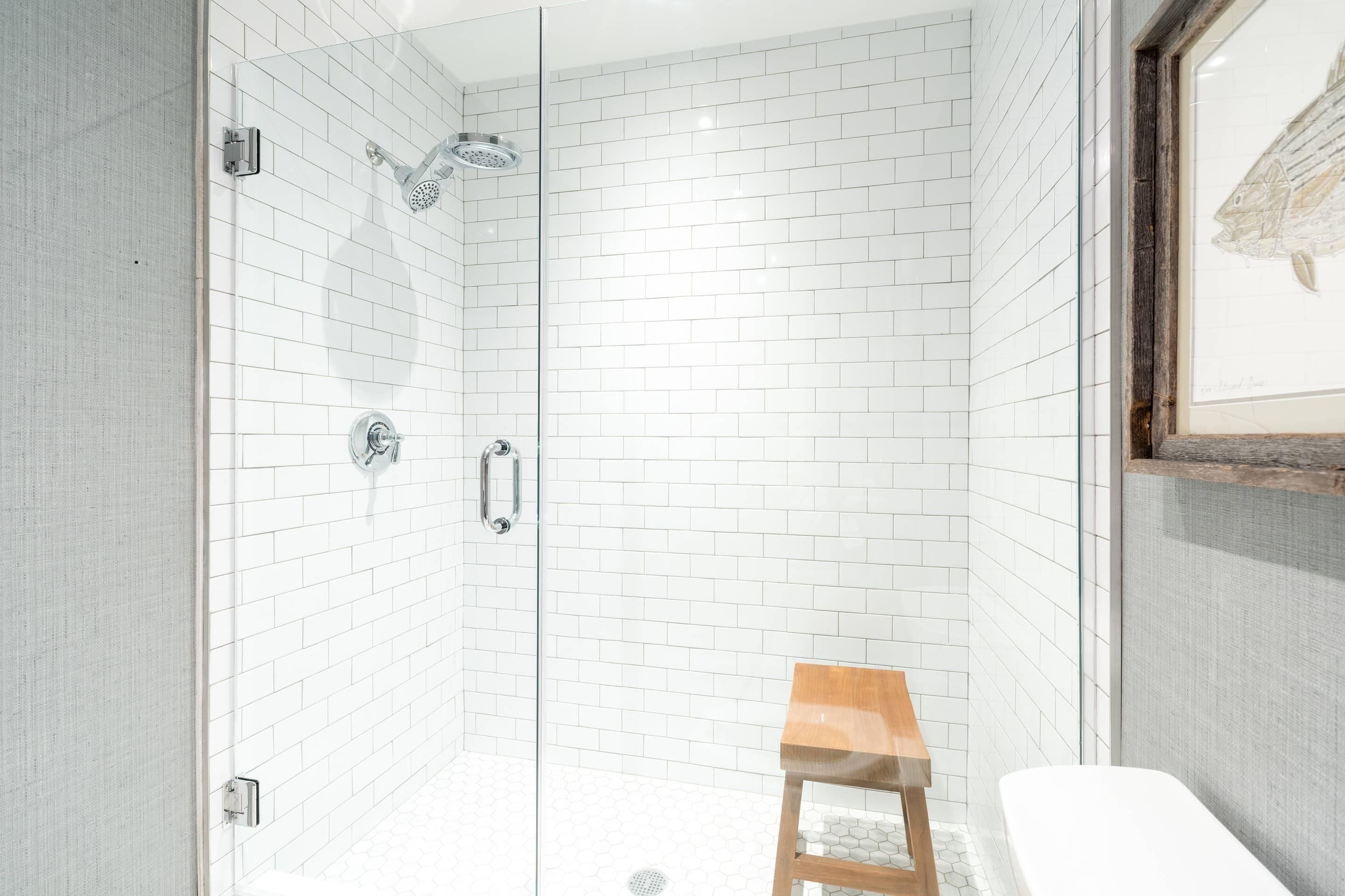 Un baño moderno con azulejos blancos tipo metro, cerramiento de ducha de vidrio, un taburete de madera y una obra enmarcada en la pared.