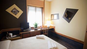 Habitación de hotel con cama, escritorio con televisor, silla, lámpara y ventana con cortinas.