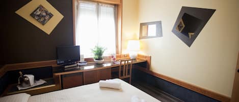 Habitación de hotel con cama, escritorio con televisor, silla, lámpara y ventana con cortinas.